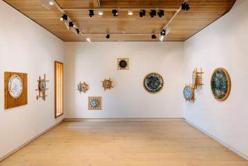 An exhibition space inside Toi MAHARA with white walls and hardwood floors circular woven artworks on display. 