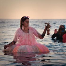 3 people standing waist deep in the ocean, surrounded by rocks peeking out of the water. One wears a wetsuit and diving gear with a catch bag, another has no shirt and a mullet, and the other is wearing a pink tulle dress with pink accessories, and holds 