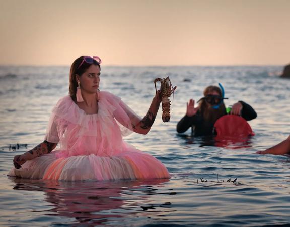 3 people standing waist deep in the ocean, surrounded by rocks peeking out of the water. One wears a wetsuit and diving gear with a catch bag, another has no shirt and a mullet, and the other is wearing a pink tulle dress with pink accessories, and holds 