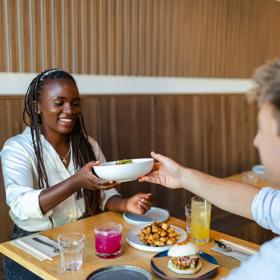 A person hands a dish to their friend across a small table during a meal.