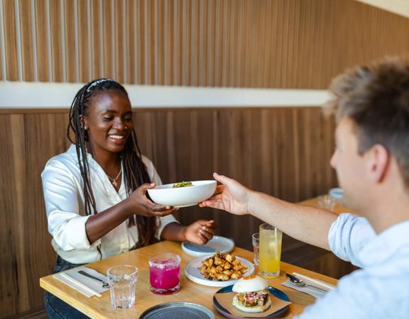 A person hands a dish to their friend across a small table during a meal.