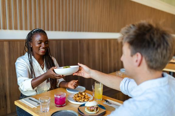 A person hands a dish to their friend across a small table during a meal.