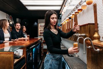 A bartender pours a pint of craft beer at Garage Project Taproom as three patrons stand at the counter.