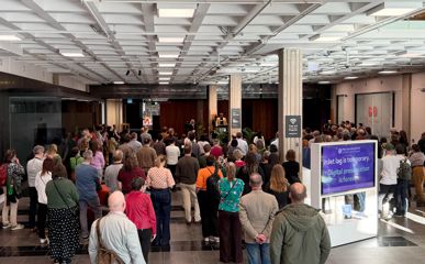 A large crowd stands in an open indoor atrium, facing a speaker during an event, with signage and display screens visible nearby.