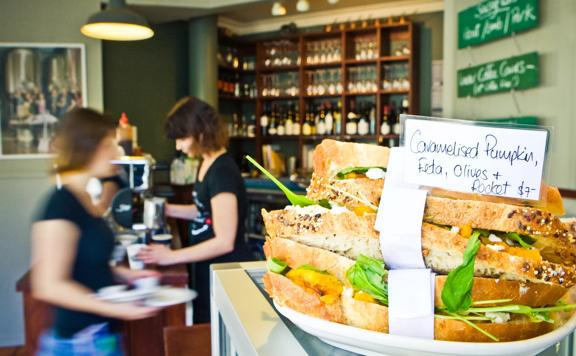 On the counter at The Larder in Miramar sits a stack of caramelised pumpkin, feta, olives, and rocket sandwiches. Staff in the background carry plates and make coffee.