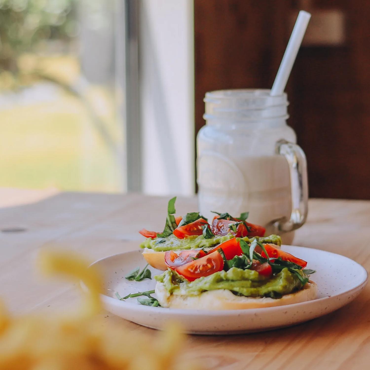 An avocado toast with quartered cherry tomato and shredded basil on a small white place and a white frothy drink in a glass mason jar on a wooden bench at Gt Fixed Bicycle Cafe in Porirua. 