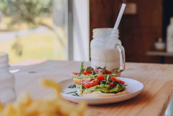 An avocado toast with quartered cherry tomato and shredded basil on a small white place and a white frothy drink in a glass mason jar on a wooden bench at Gt Fixed Bicycle Cafe in Porirua.