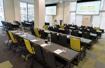 A conference room set up for a presentation with five rows of tables and chairs facing a podium and two projector screens suspended from the ceiling on either side.