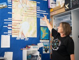 A person wearing a House of Science T-shirt gestures to a map on the classroom wall.