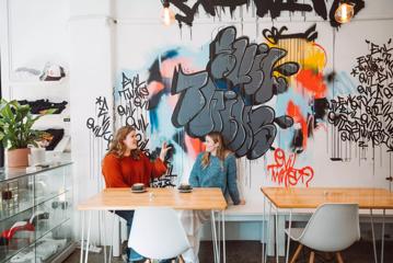 Two friends sit and enjoy coffee at Evil Twins Coffee, a café in Te Aro, Wellington. The bright space has graffiti on the wall, wooden tables and concrete floors.