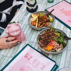 A close-up of two dishes, two menus, and two cocktails in animal-shaped cups on a pale green table at Mr. Go's.