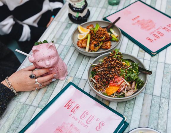 A close-up of two dishes, two menus, and two cocktails in animal-shaped cups on a pale green table at Mr. Go's.