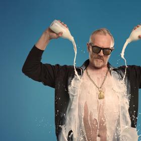 Greg Davies in an open black shirt pouring two bottles of milk on himself.
