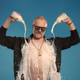 Greg Davies in an open black shirt pouring two bottles of milk on himself.