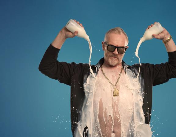 Greg Davies in an open black shirt pouring two bottles of milk on himself.
