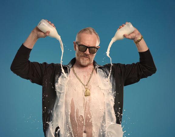 Greg Davies in an open black shirt pouring two bottles of milk on himself.