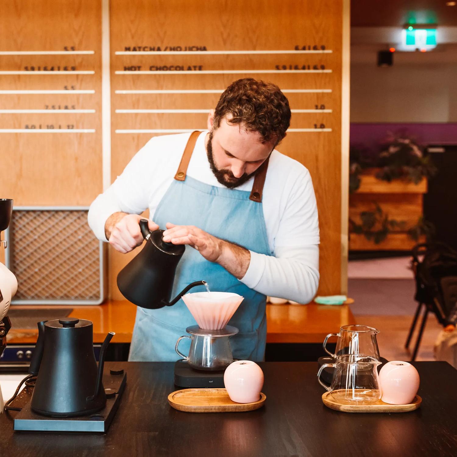 A barista in a blue apron prepares pour-over coffee.