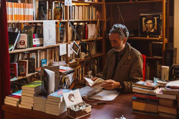 A person sits behind a desk looking at a book, while surrounded by piles and shelves of books in The Undercurrent bookstore.