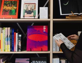 Light beige shelves with various books about contemporary art. A person's arms come into the right of the frame, wearing black sleeves holding a small white book with the title "Territory Unknown" in black all-caps serif font.