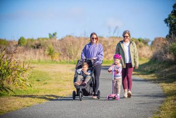 Two adults walking along Te Ara o Whareroa, a sealed pathway through Queen Elizabeth Park, one is pushing a small child in a push chair, and another small child scooters ahead of the group.