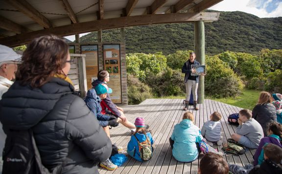 A group of school-aged children sit on a wooden deck listening to a talk about native birds on Kapiti Island.