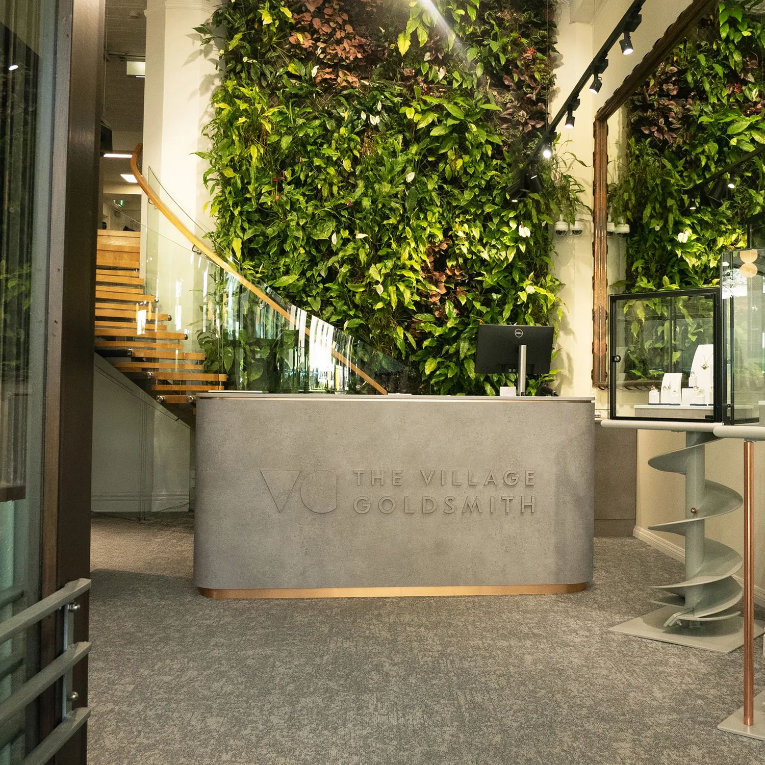 Reception area at The Village Goldsmith, featuring a living green wall, display cases, and a glass-railed staircase.