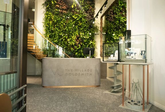 Reception area at The Village Goldsmith, featuring a living green wall, display cases, and a glass-railed staircase.