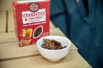 A box of upcycled grain project grain crisps sit on a cardboard box next to a white bowl filled with crisps.