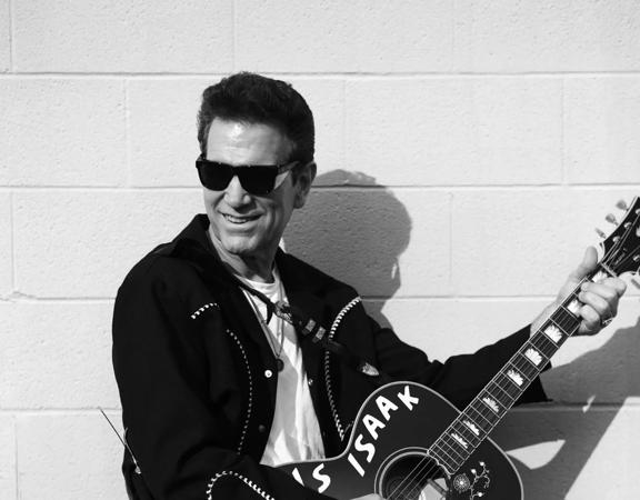 Chris Isaak standing in front of a white brick wall, wearing a black leather jacket and sunglasses playing a guitar.