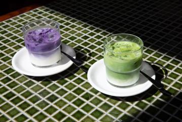 Two glasses sit on white plates on a green tile table. The drinks are milky and one is purple, the other is green.