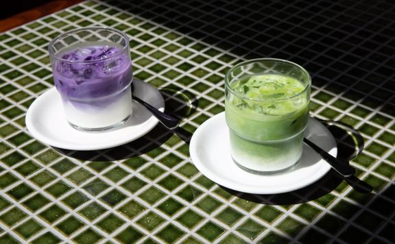 Two glasses sit on white plates on a green tile table. The drinks are milky and one is purple, the other is green.