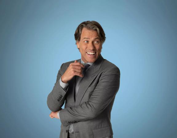 John Bishop, wearing a grey suit, smiles and poses in front of a light blue wall. 