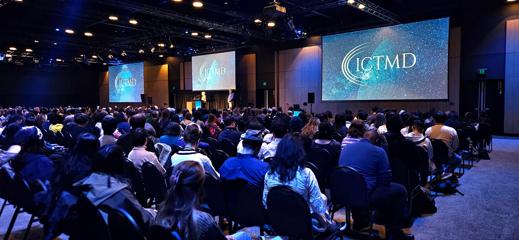 People sitting on rows of chairs in a large conference space, facing towards projector screens that have the ICTMD logo on display.
