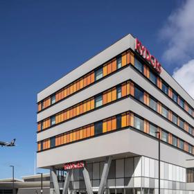 The exterior of Rydges Hotel at Wellington Airport is a grey building with orange window shades. A black Air New Zealand plane takes off in the distance.