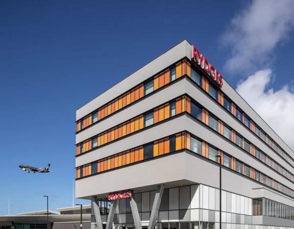 The exterior of Rydges Hotel at Wellington Airport is a grey building with orange window shades. A black Air New Zealand plane takes off in the distance.