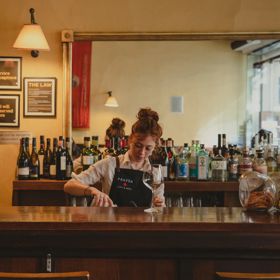 Person working behind a wooden bar lined with bottles and glassware in a warm, cozy café or cocktail bar setting.