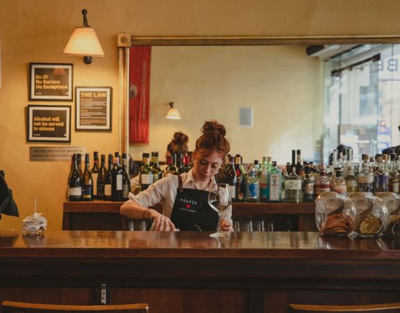 Person working behind a wooden bar lined with bottles and glassware in a warm, cozy café or cocktail bar setting.