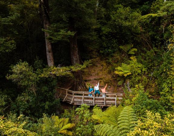 Three people are on a wooden bridge waving on Tane's Track, a walking trail in Upper Hutt, Hutt Valley. They are surrounded by lush greenery.