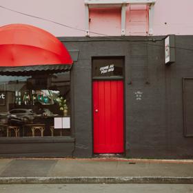The exteriror of Margot in Newtown. it is painted black with  a red door and cover over thr window. Their sign sits on the window as well as a smell decal on the door that says Come as you are.
