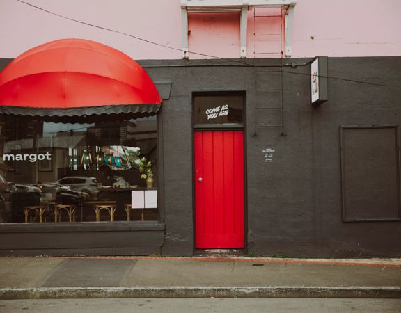 The exteriror of Margot in Newtown. it is painted black with  a red door and cover over thr window. Their sign sits on the window as well as a smell decal on the door that says Come as you are.