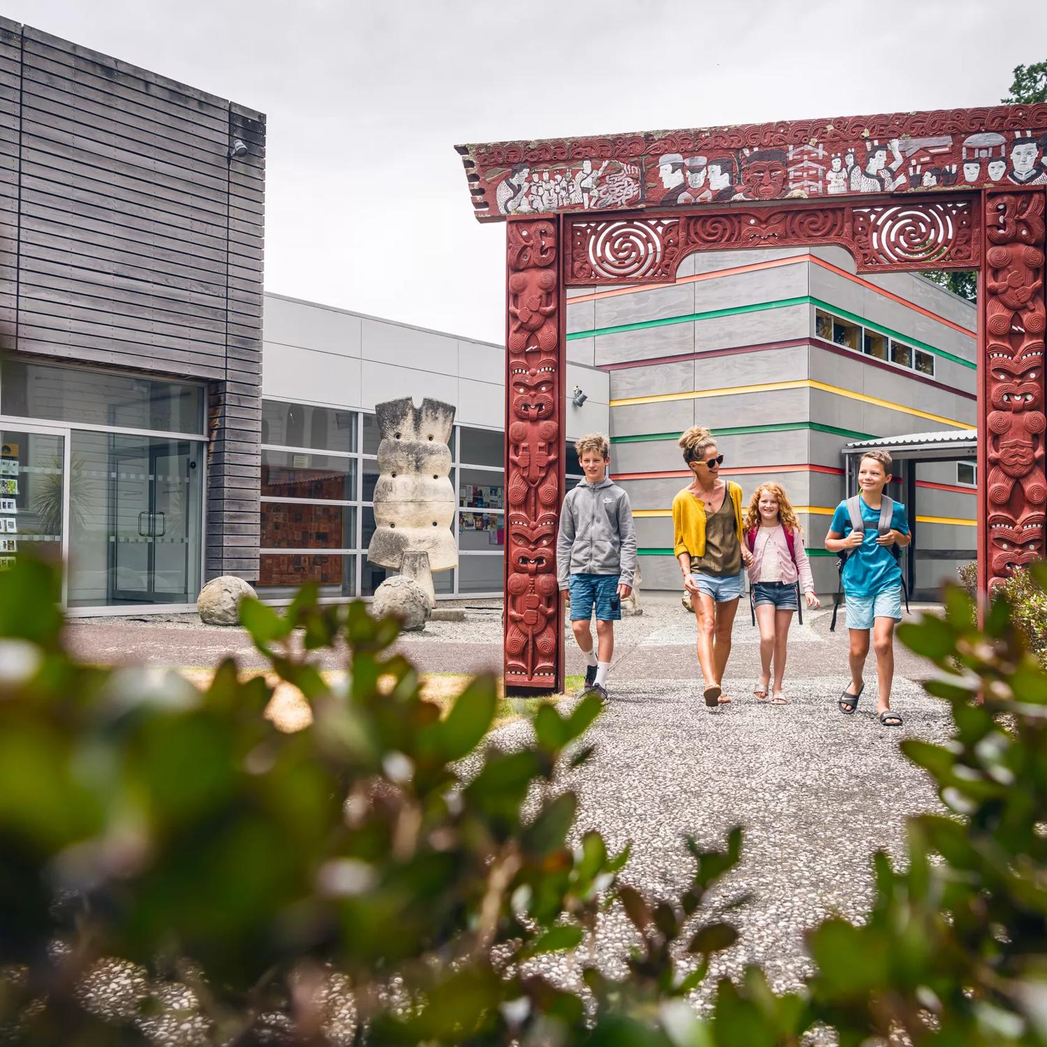 The forecourt outside Aratoi. There is a large archway with carved Māori figures and motifs in the middle of the forecourt. A family of three are walking through it. The Aratoi buildings are in the background.