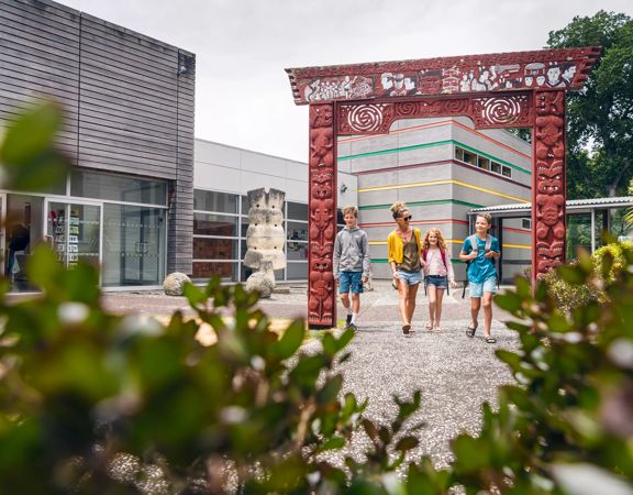 The forecourt outside Aratoi. There is a large archway with carved Māori figures and motifs in the middle of the forecourt. A family of three are walking through it. The Aratoi buildings are in the background.