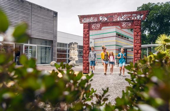 The forecourt outside Aratoi. There is a large archway with carved Māori figures and motifs in the middle of the forecourt. A family of three are walking through it. The Aratoi buildings are in the background.