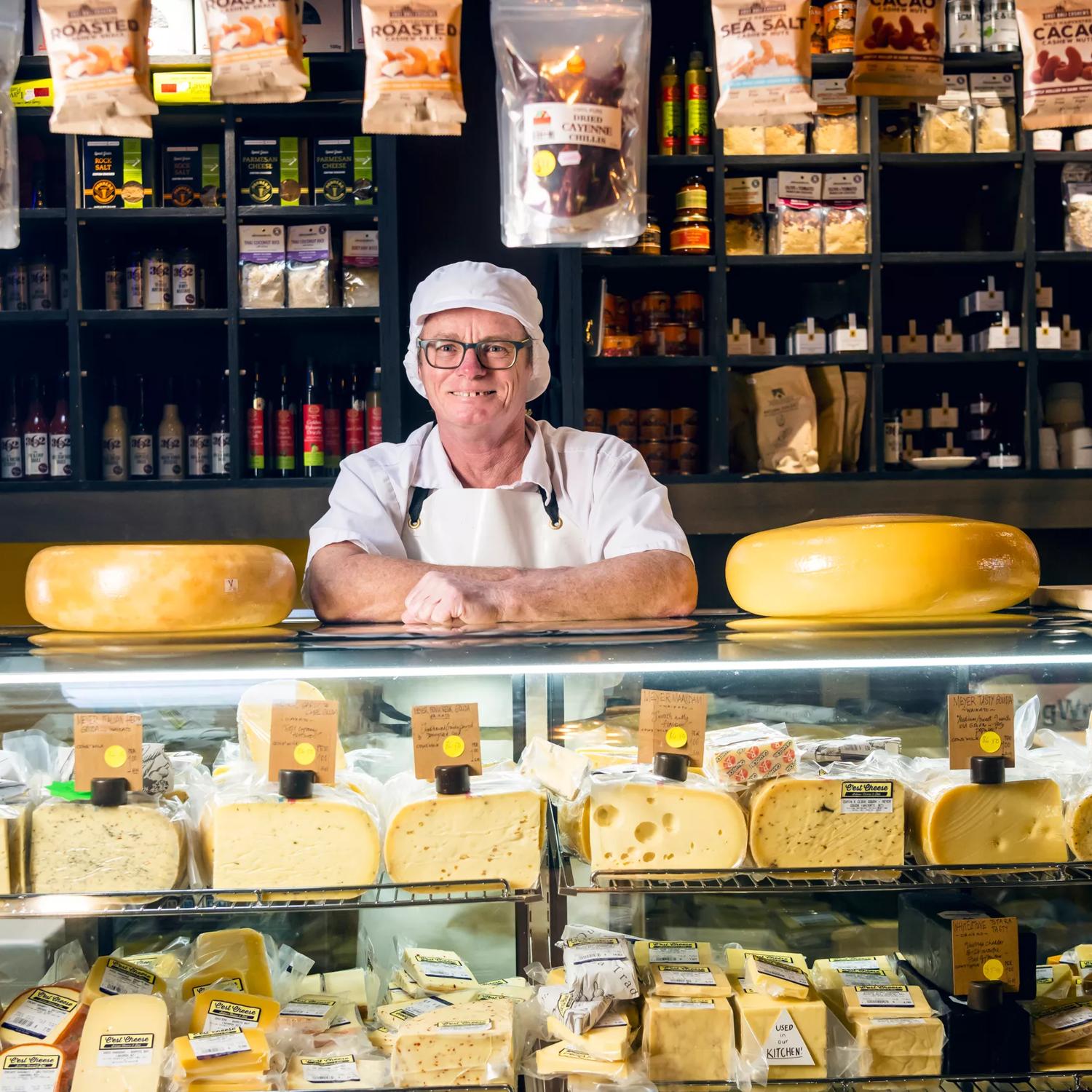 A smiling cheesemaker wearing a white uniform is behind the counter at C'est Cheese, an artisanal deli in Featherston, Wairarapa.