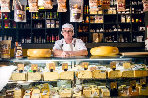 A smiling cheesemaker wearing a white uniform is behind the counter at C'est Cheese, an artisanal deli in Featherston, Wairarapa.