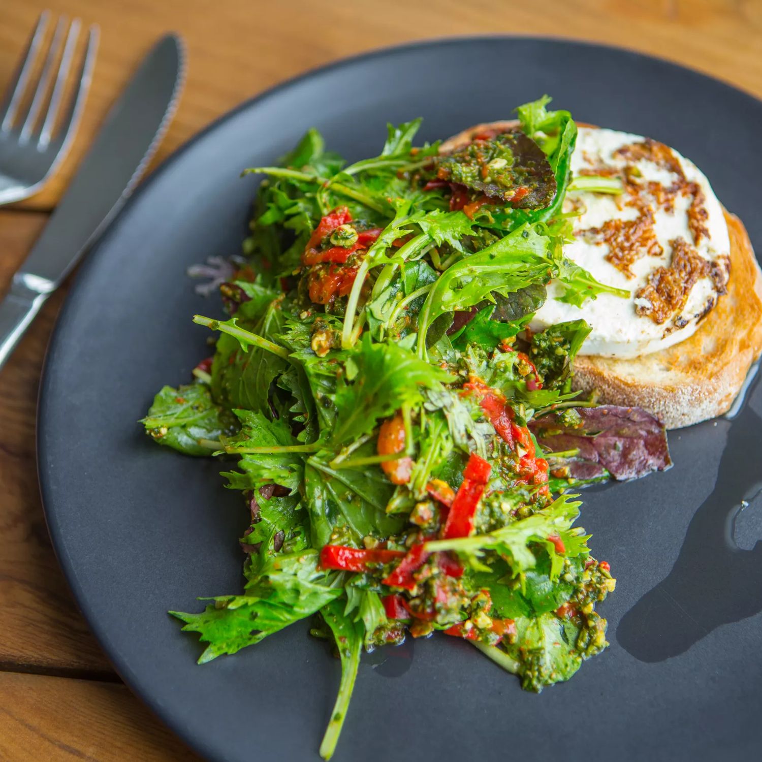 Fresh green salad with red chilies and a poached egg on toasted bread, served on a dark plate with fork and knife.