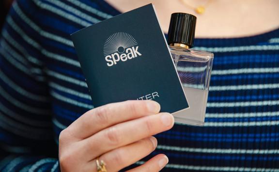 A person is holding a booklet and perfume bottle from Speak Scents, a New Zealand-based fragrance company. 