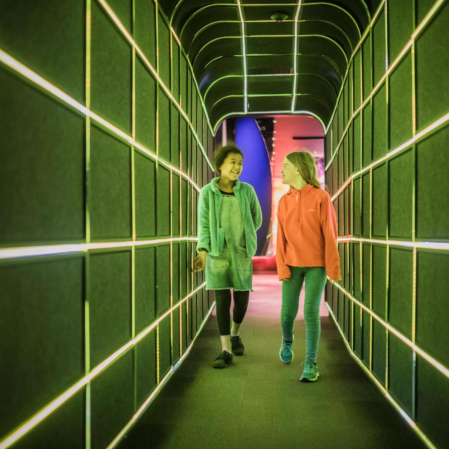 2 children walking through a tunnel at Space place lit up by green LED lights.