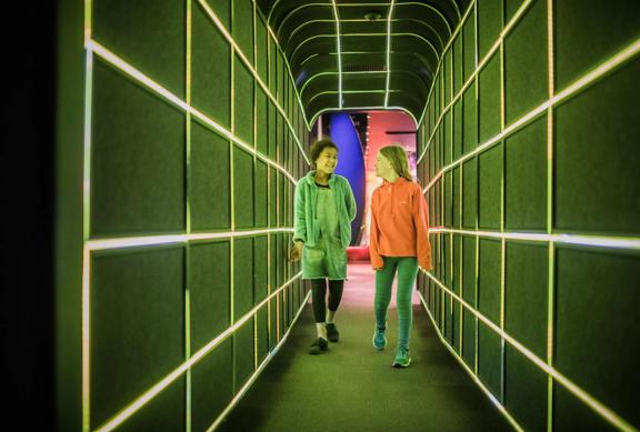 2 children walking through a tunnel at Space place lit up by green LED lights.