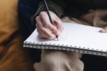 A person using a pencil to draw in a sketchbook on their lap.